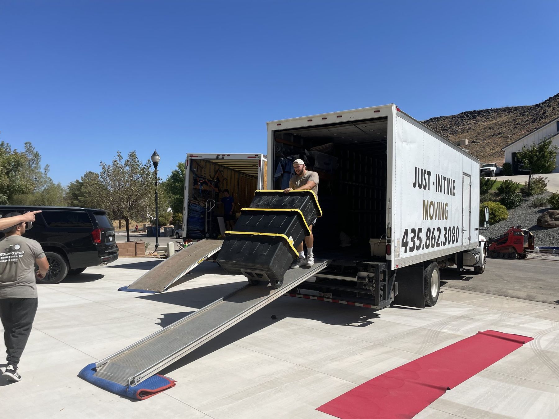 A Just-In Time truck with the loading ramp down and a red floor runner protecting the driveway, red rock hills behind
