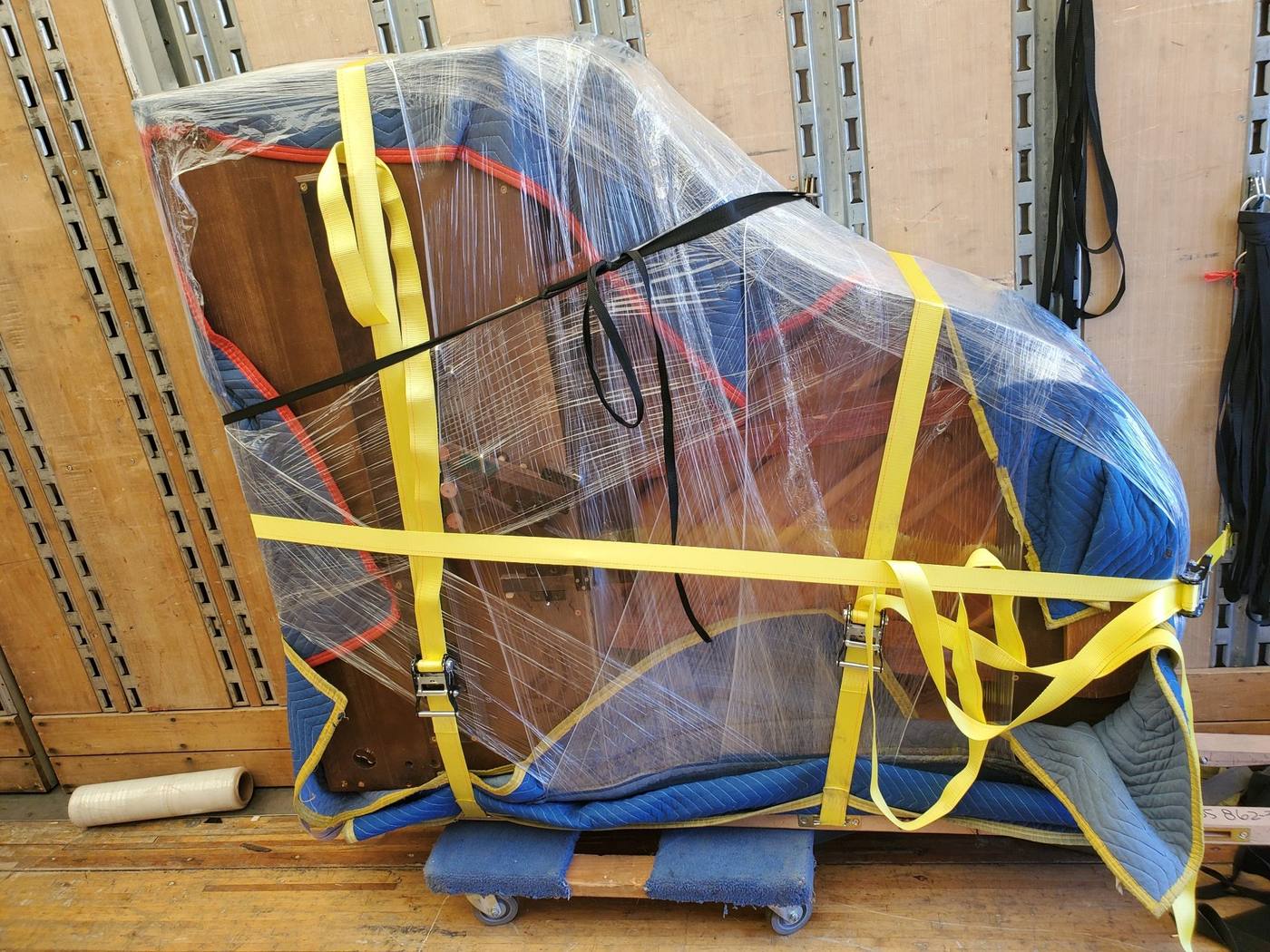 A piano shrink-wrapped, padded and secured with yellow ratchet straps on a moving dolly