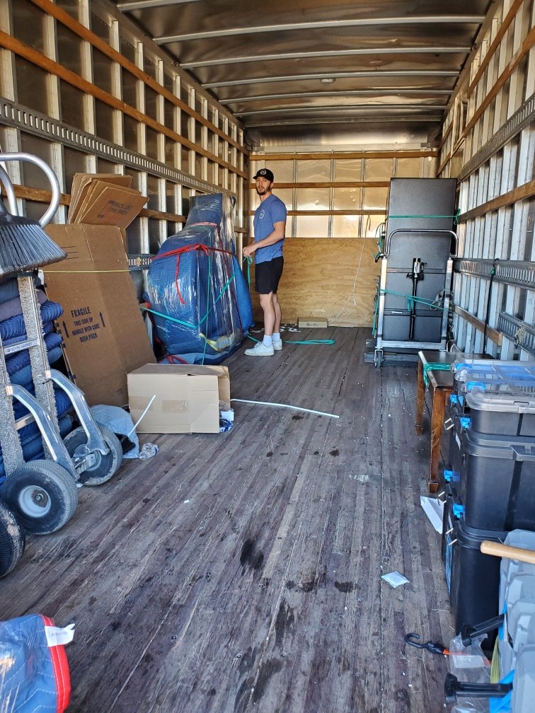 The long interior of a moving truck with dollies and equipment, a crew member working at the far end